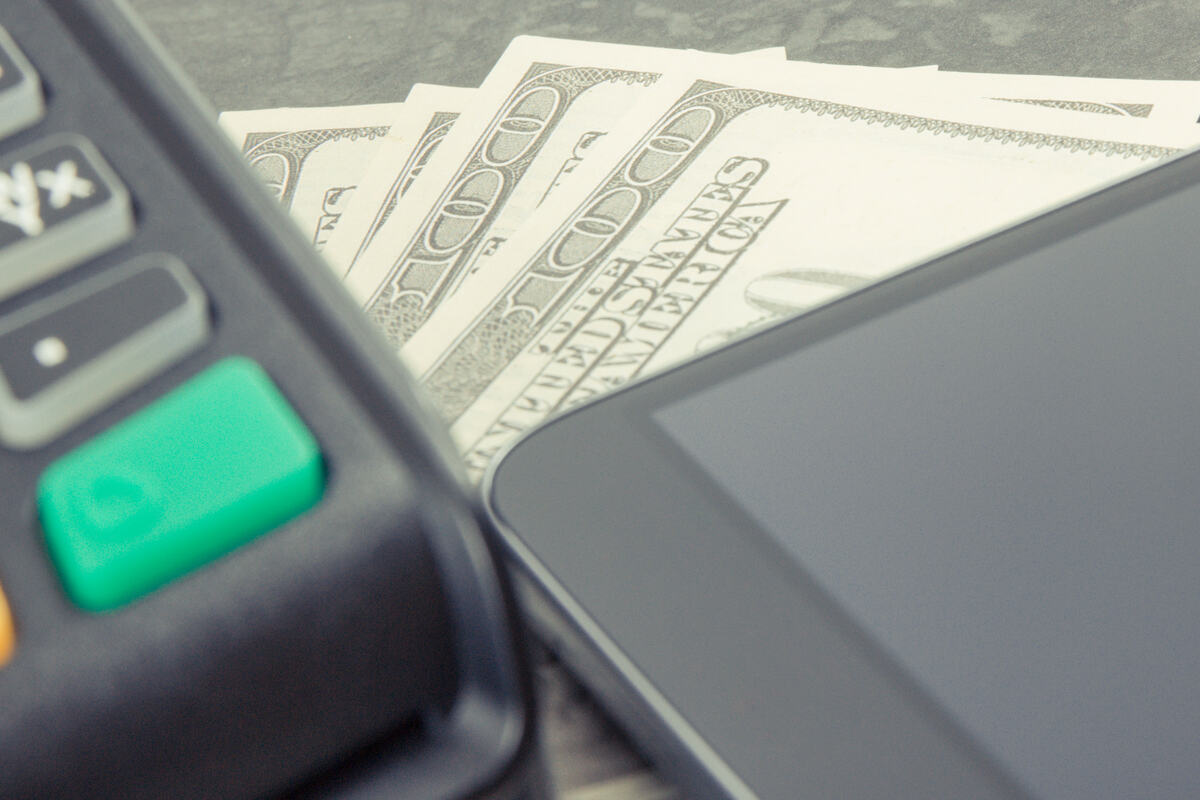 Close-up of a payment terminal, smartphone, and $100 bills showing digital and cash transactions.